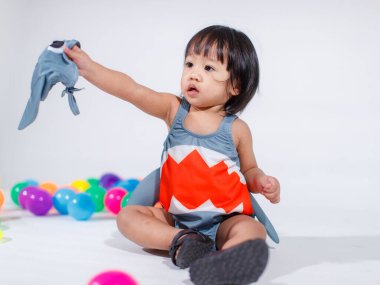 Renkli plastik toplar ve beyaz arka planda yüzük oyuncakları ile yerde oturan şirin gri köpek balığı kostümlü Asyalı küçük kız bebek modelinin stüdyo fotoğrafı.