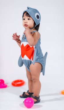Renkli plastik toplar ve beyaz arka planda yüzük oyuncaklarıyla yerde duran şirin gri köpek balığı kostümlü Asyalı kız bebek modelinin stüdyo fotoğrafı..
