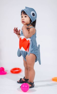 Renkli plastik toplar ve beyaz arka planda yüzük oyuncaklarıyla yerde duran şirin gri köpek balığı kostümlü Asyalı kız bebek modelinin stüdyo fotoğrafı..