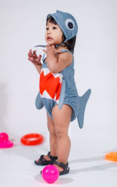 Renkli plastik toplar ve beyaz arka planda yüzük oyuncaklarıyla yerde duran şirin gri köpek balığı kostümlü Asyalı kız bebek modelinin stüdyo fotoğrafı..