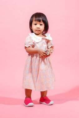 Portre stüdyosu küçük tatlı Asyalı anaokulu anaokulu kızı kız modelini gündelik uzun elbiseler ve ayakkabılar içinde elinde pembe arka planda yüz dolarlık banknotla kameraya bakarken çekti..