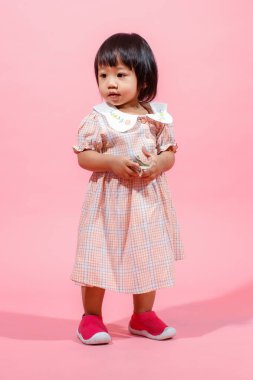 Portre stüdyosu küçük tatlı Asyalı anaokulu anaokulu kızı kız modelini gündelik uzun elbiseler ve ayakkabılar içinde elinde pembe arka planda yüz dolarlık banknotla kameraya bakarken çekti..