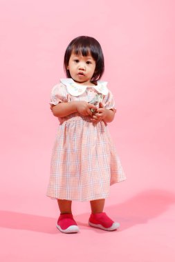 Portre stüdyosu küçük tatlı Asyalı anaokulu anaokulu kızı kız modelini gündelik uzun elbiseler ve ayakkabılar içinde elinde pembe arka planda yüz dolarlık banknotla kameraya bakarken çekti..