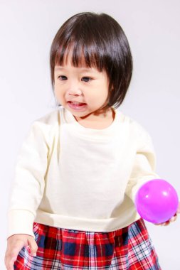 Küçük, şirin, siyah saçlı, Asyalı, ekoseli etekli ve gülümseyen beyaz arka planda tek başına renkli yuvarlak toplarla oynayan küçük bir kızın stüdyo fotoğrafı..