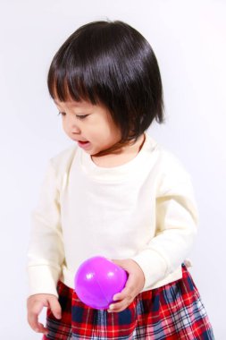 Küçük, şirin, siyah saçlı, Asyalı, ekoseli etekli ve gülümseyen beyaz arka planda tek başına renkli yuvarlak toplarla oynayan küçük bir kızın stüdyo fotoğrafı..