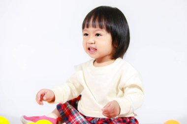 Küçük, şirin, kısa, siyah saçlı, Asyalı, ekoseli etekli, gülen, beyaz arka planda tek başına renkli yuvarlak toplarla oynayan küçük kız modelin stüdyo fotoğrafı..