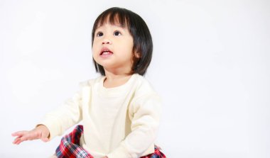 Küçük, şirin, kısa, siyah saçlı, Asyalı, ekose etekli, beyaz arka planda oturan ve gülümseyen kız çocuğunun stüdyo fotoğrafı.