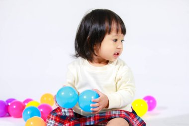 Küçük, şirin, kısa, siyah saçlı, Asyalı, ekoseli etekli, gülen, beyaz arka planda tek başına renkli yuvarlak toplarla oynayan küçük kız modelin stüdyo fotoğrafı..