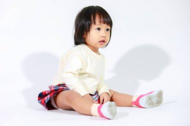 Küçük, şirin, kısa, siyah saçlı, Asyalı, ekose etekli, beyaz arka planda oturan ve gülümseyen kız çocuğunun stüdyo fotoğrafı.