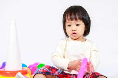 Küçük şirin Asyalı kız bebek modelin stüdyo fotoğrafı. Günlük kıyafetleriyle yerde oturmuş renkli plastik yüzüklerle oynuyorlar. İnşaat piramidi oyuncakları ve beyaz arka planda küçük toplar..