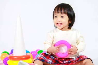 Küçük şirin Asyalı kız bebek modelin stüdyo fotoğrafı. Günlük kıyafetleriyle yerde oturmuş renkli plastik yüzüklerle oynuyorlar. İnşaat piramidi oyuncakları ve beyaz arka planda küçük toplar..
