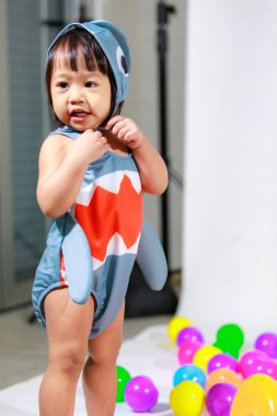 Renkli plastik toplar ve beyaz arka planda yüzük oyuncaklarıyla yerde duran şirin gri köpek balığı kostümlü Asyalı kız bebek modelinin stüdyo fotoğrafı..