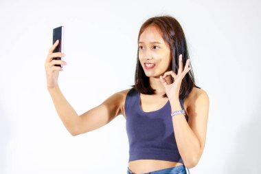 Asyalı genç kız modelin izole edilmiş bir fotoğrafı. Üstü açık tişört ve kot pantolon içinde. İnternette akıllı telefon görüntüsü var..