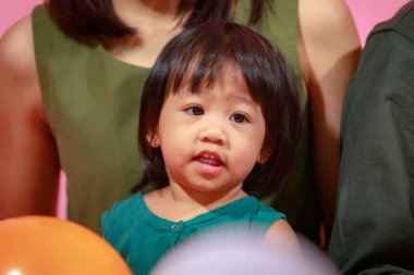 Genç mutlu Asyalı aile babasının, annesinin ve küçük tatlı kızının birlikte yerde oturup pembe arka planda renkli balonlar tutarak kameraya gülümserkenki portresi..