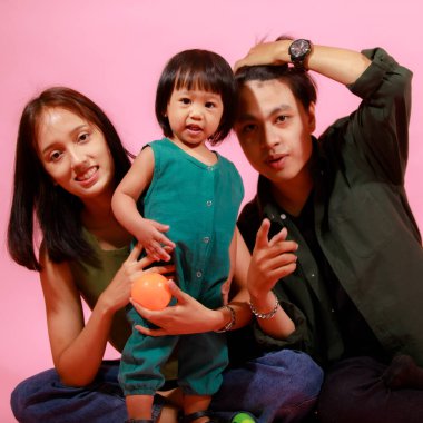 Genç mutlu Asyalı aile babasının, annesinin ve küçük tatlı kızının birlikte yerde oturup pembe arka planda renkli balonlar tutarak kameraya gülümserkenki portresi..