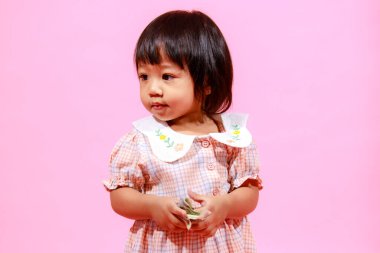 Portre stüdyosu küçük tatlı Asyalı anaokulu anaokulu kızı kız modelini gündelik uzun elbiseler ve ayakkabılar içinde elinde pembe arka planda yüz dolarlık banknotla kameraya bakarken çekti..