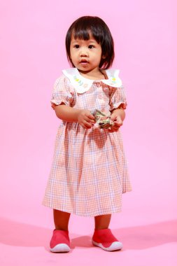 Portre stüdyosu küçük tatlı Asyalı anaokulu anaokulu kızı kız modelini gündelik uzun elbiseler ve ayakkabılar içinde elinde pembe arka planda yüz dolarlık banknotla kameraya bakarken çekti..