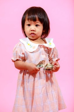 Portre stüdyosu küçük tatlı Asyalı anaokulu anaokulu kızı kız modelini gündelik uzun elbiseler ve ayakkabılar içinde elinde pembe arka planda yüz dolarlık banknotla kameraya bakarken çekti..