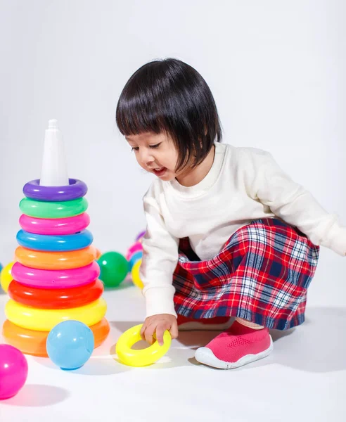 Küçük şirin Asyalı kız bebek modelin stüdyo fotoğrafı. Günlük kıyafetleriyle yerde oturmuş renkli plastik yüzüklerle oynuyorlar. İnşaat piramidi oyuncakları ve beyaz arka planda küçük toplar..