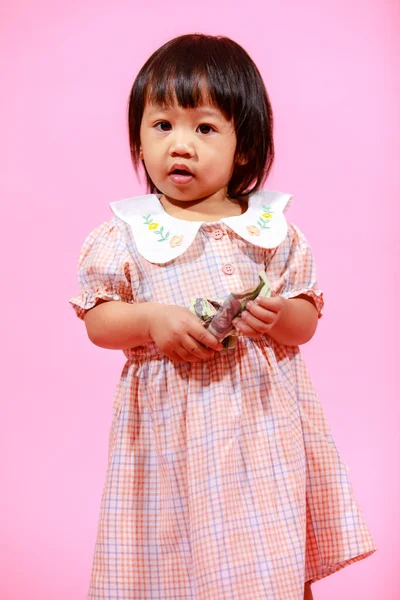 肖像工作室拍摄了可爱的亚洲幼稚园女模特儿 她穿着休闲装和鞋子 站在那里看着相机 手里拿着100美元钞票 背景是粉色的 — 图库照片
