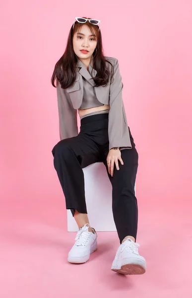 ポートレートスタジオショットのアジアの若い都市の流行の女性ヒップスターティーンエイジャーのファッションモデルでカジュアル作物トップストリート着用ジャケット保持サングラスで頭の上に座る上のピンクの背景に白い正方形のボックススツール — ストック写真