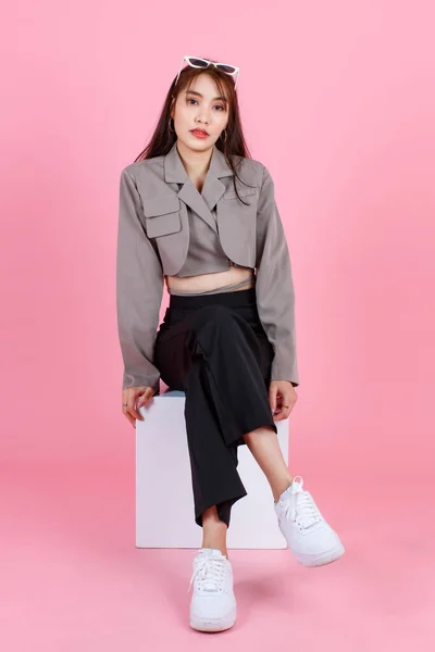 ポートレートスタジオショットのアジアの若い都市の流行の女性ヒップスターティーンエイジャーのファッションモデルでカジュアル作物トップストリート着用ジャケット保持サングラスで頭の上に座る上のピンクの背景に白い正方形のボックススツール — ストック写真