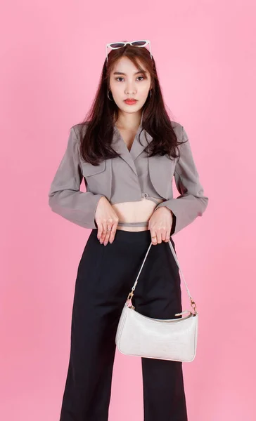 亚洲流行女嬉皮士少年模特儿穿着夹克衫太阳镜运动鞋手持手提包看着粉色背景的相机 — 图库照片