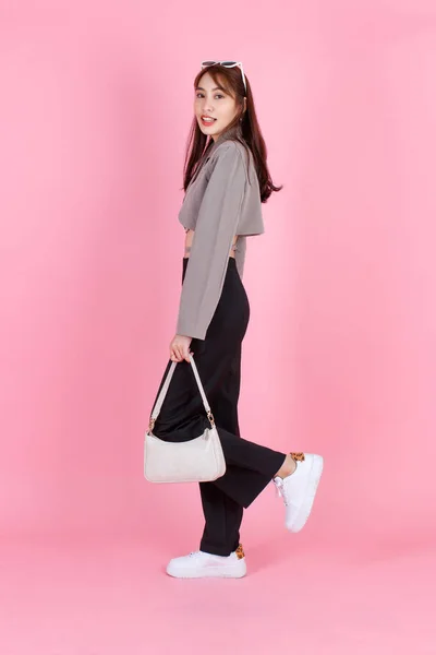 アジアの流行のファッショナブルな女性ヒップスターティーンモデルのポートレートスタジオショットカジュアルな作物トップストリートでは ピンクの背景にカメラを見て歩くハンドバッグ財布を保持ジャケットサングラススニーカーを身に着けています — ストック写真