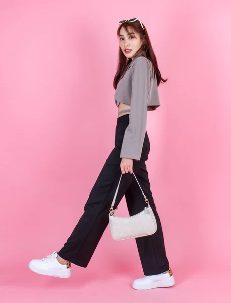 亚洲流行女嬉皮士少年模特儿穿着夹克衫太阳镜运动鞋提着手提包走在粉色背景下看着相机的肖像工作室照片 — 图库照片