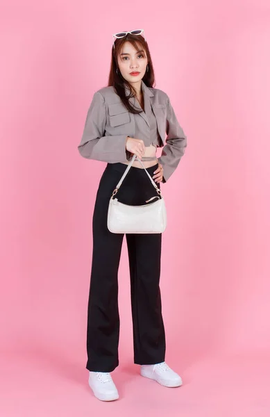 亚洲流行女嬉皮士少年模特儿穿着夹克衫太阳镜运动鞋手持手提包看着粉色背景的相机 — 图库照片