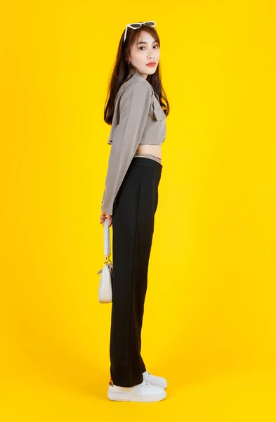 アジアの流行のファッショナブルな女性ヒップスターティーンモデルのポートレートスタジオショットカジュアルな作物トップストリートでは 黄色の背景にカメラを見てハンドバッグ財布を保持ジャケットサングラススニーカーを身に着けています — ストック写真