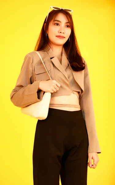 アジアの流行のファッショナブルな女性ヒップスターティーンモデルのポートレートスタジオショットカジュアルな作物トップストリートでは 黄色の背景にカメラを見てハンドバッグ財布を保持ジャケットサングラススニーカーを身に着けています — ストック写真