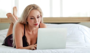 Beyaz kadın, sarışın, siyah seksi iç çamaşırı giyen seksi kadın modelin yakın çekimleri yatak odasındaki dizüstü bilgisayarla internette gezinirken gülümseyerek uzandı.