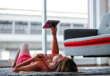 Beyaz kadın, sarışın, seksi manken, pembe seksi iç çamaşırlı, halı döşemesine uzanmış, bacakları tablet bilgisayarın üzerine koyup reklam alanı fotokopisi çektiriyor.