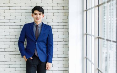 Portre stüdyosu Asyalı yakışıklı, profesyonel, başarılı, iş adamı, resmi mavi kravat takmış, gülümseyen bir şekilde ofis penceresinin yanındaki beyaz tuğla duvardaki kameraya baktı.