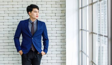 Portre stüdyosu Asyalı yakışıklı, profesyonel, başarılı, iş adamı, resmi mavi kravat takmış, gülümseyen bir şekilde ofis penceresinin yanındaki beyaz tuğla duvardaki kameraya baktı.