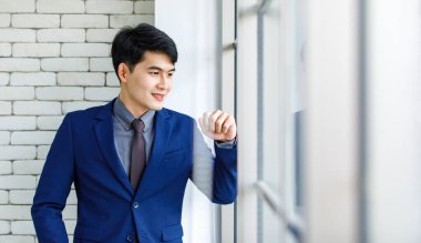 Portre stüdyosu Asyalı yakışıklı, profesyonel, başarılı, iş adamı, resmi mavi kravat takmış, gülümseyen bir şekilde ofis penceresinin yanındaki beyaz tuğla duvardaki kameraya baktı.