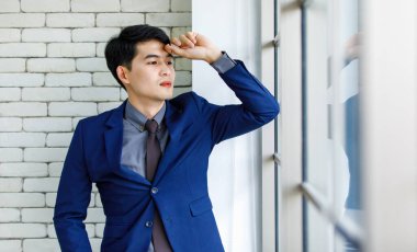 Portre stüdyosu Asyalı yakışıklı, profesyonel, başarılı, iş adamı, resmi mavi kravat takmış, gülümseyen bir şekilde ofis penceresinin yanındaki beyaz tuğla duvardaki kameraya baktı.