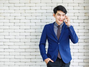 Asyalı yakışıklı, profesyonel, başarılı, iş adamı, resmi mavi kravatlı, elinde akıllı telefonuyla beyaz duvar arkasında gülümseyen bir adamın portresi..