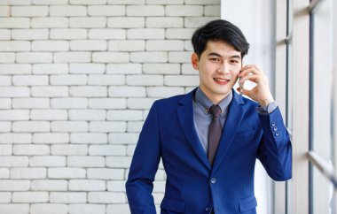 Asyalı yakışıklı, profesyonel, başarılı, iş adamı, resmi mavi kravatlı, elinde akıllı telefonuyla beyaz duvar arkasında gülümseyen bir adamın portresi..