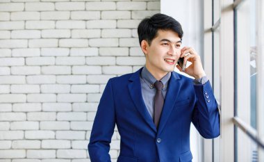 Asyalı yakışıklı, profesyonel, başarılı, iş adamı, resmi mavi kravatlı, elinde akıllı telefonuyla beyaz duvar arkasında gülümseyen bir adamın portresi..