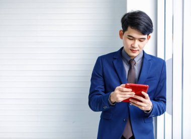 Milenyum Asyalı yakışıklı, kendine güvenen, profesyonel başarılı, erkek, yönetici, iş adamı, resmi mavi takım elbiseli, elinde akıllı telefonuyla gökdelen ofis penceresinin yanında duran gülümseyen adam.