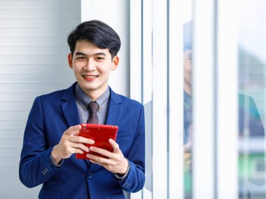 Milenyum Asyalı yakışıklı, kendine güvenen, profesyonel başarılı, erkek, yönetici, iş adamı, resmi mavi takım elbiseli, elinde akıllı telefonuyla gökdelen ofis penceresinin yanında duran gülümseyen adam.