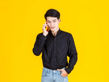 Milenyum Asyalı heyecanlı iş adamı modelinin atölye fotoğrafı. Uzun kollu günlük siyah gömlekli ve kot pantolonlu sarı arka plandaki akıllı telefonu arıyor..