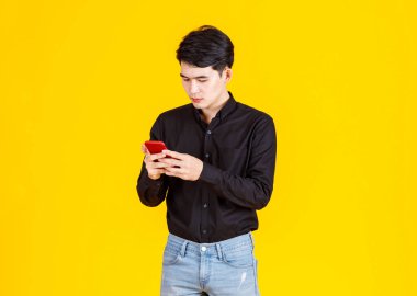 Milenyum Asyalı heyecanlı erkek iş adamı modelinin atölye fotoğrafı. Uzun kollu günlük siyah gömlekli ve kot pantolonlu. Elinde kırmızı akıllı telefon, sarı arka planda sanal video oyunu oynuyor..