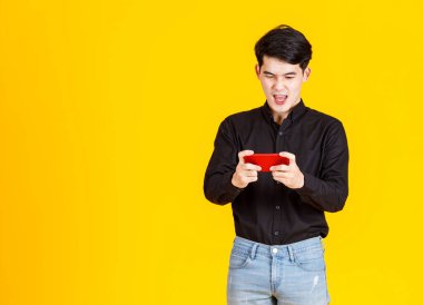 Milenyum Asyalı heyecanlı erkek iş adamı modelinin atölye fotoğrafı. Uzun kollu günlük siyah gömlekli ve kot pantolonlu. Elinde kırmızı akıllı telefon, sarı arka planda sanal video oyunu oynuyor..