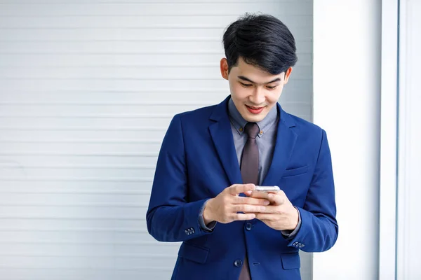 千年のアジアのハンサムなスマート自信を持ってプロの成功の男性経営者ビジネスマンの起業家の正式な青いスーツの笑顔超高層ビルのオフィスの窓の近くに立っているスマートフォンを保持 — ストック写真