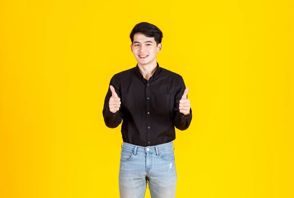 ポートレートスタジオショットの千年のアジアのプロの成功した男性ビジネスマンでカジュアルな黒シャツ立って手でジーンズポケットに親指を見上げ笑顔カメラで黄色の背景 — ストック写真