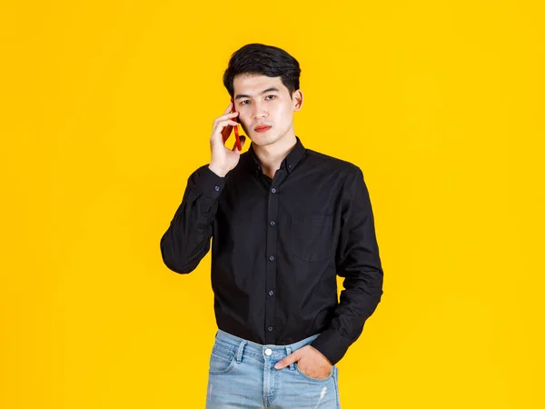 スタジオショットの千年のアジアの興奮男性ビジネスマンモデルでカジュアルな黒の長袖シャツとジーンズスタンド黄色の背景にスマートフォンを呼び出します — ストック写真