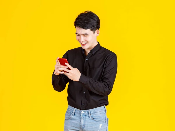 スタジオショットの千年のアジアの興奮男性ビジネスマンモデルでカジュアルな黒の長袖シャツとジーンズ立って手で赤いスマートフォンを保持黄色の背景に仮想ビデオゲームをプレイ — ストック写真